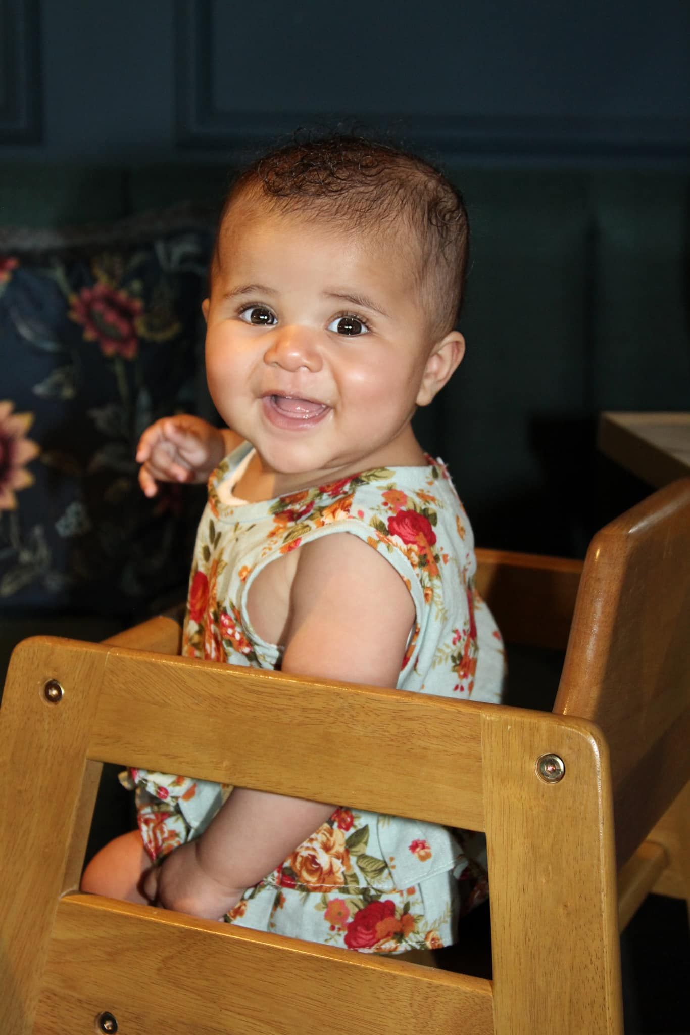 Smiling baby in a floral dress sitting in a wooden high chair.