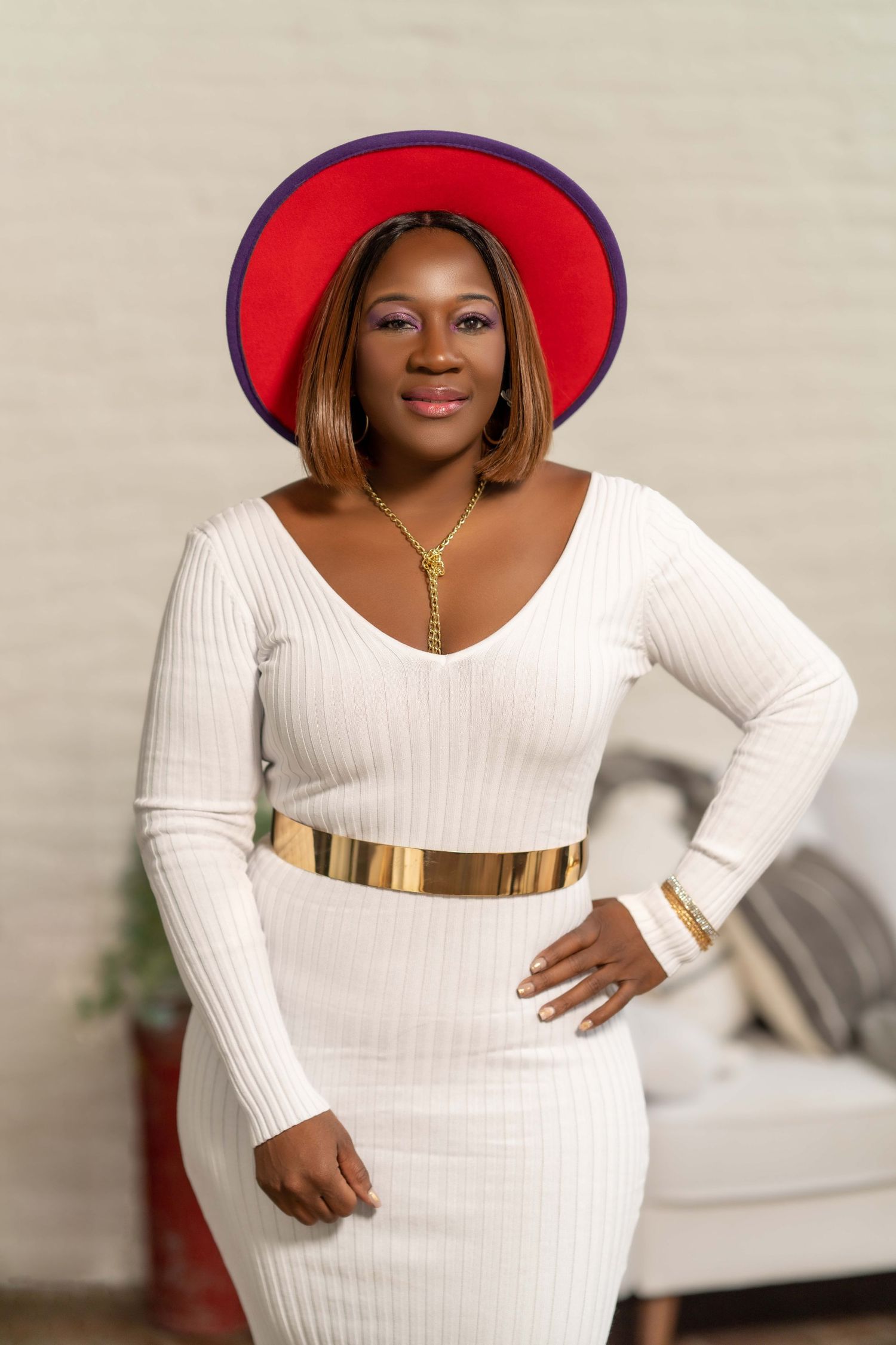 Stylish portrait of a woman in a white ribbed dress with gold belt and red hat against neutral background.