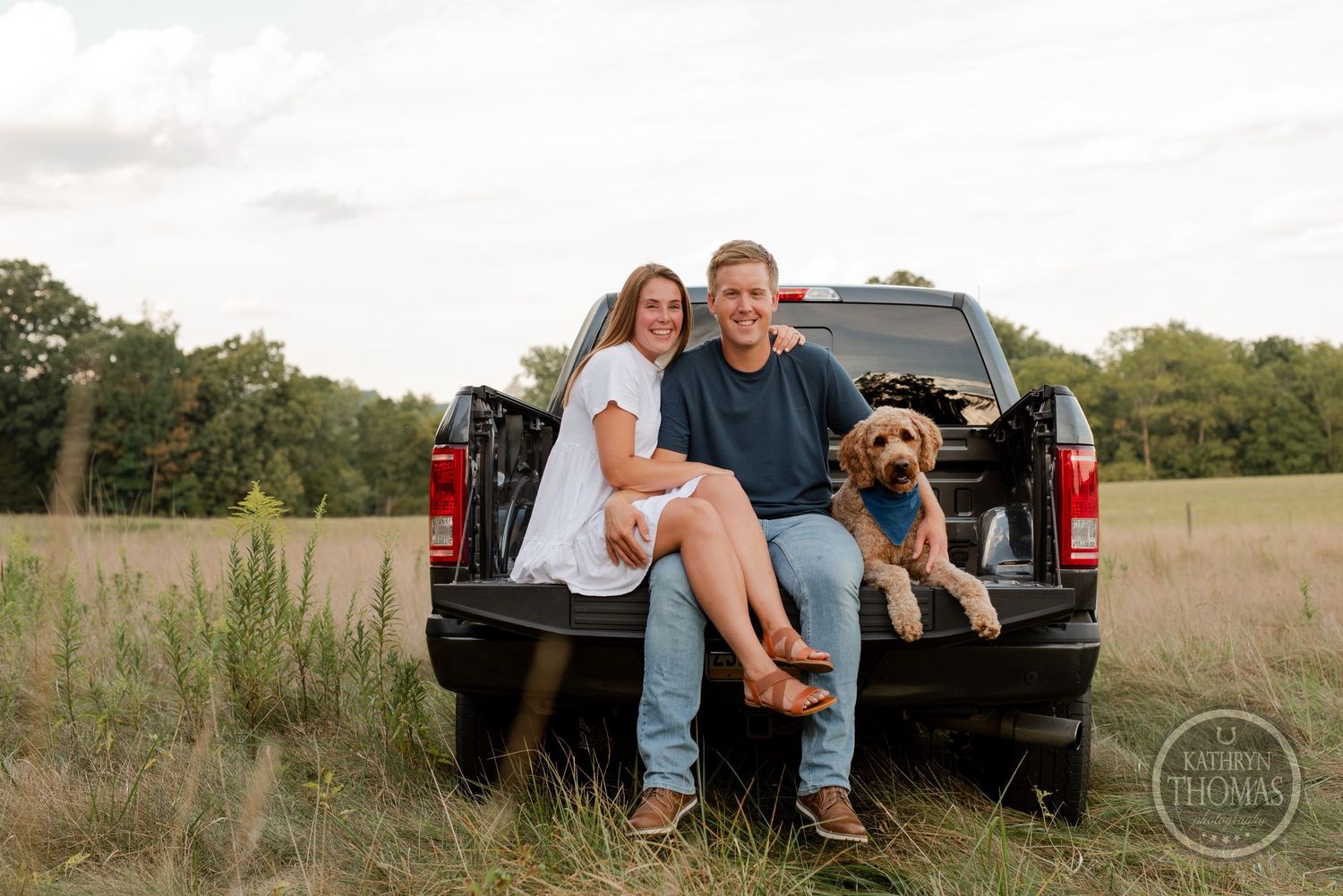 A couple sits on pickup truck tailgate with their goldendoodle in rustic field setting.