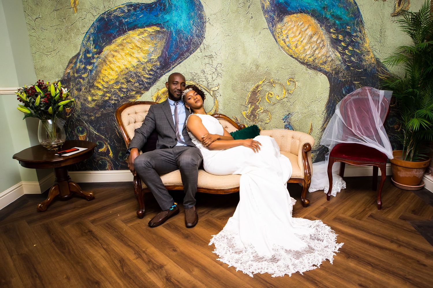 Elegant wedding couples seated on a vintage sofa with a vibrant peacock mural background recreate a stunning and artistic scene.
