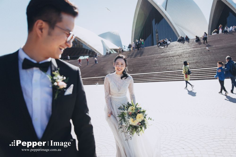 Smiling bride with bouquet and groom in tuxedo posing at Sydney Opera House plaza.