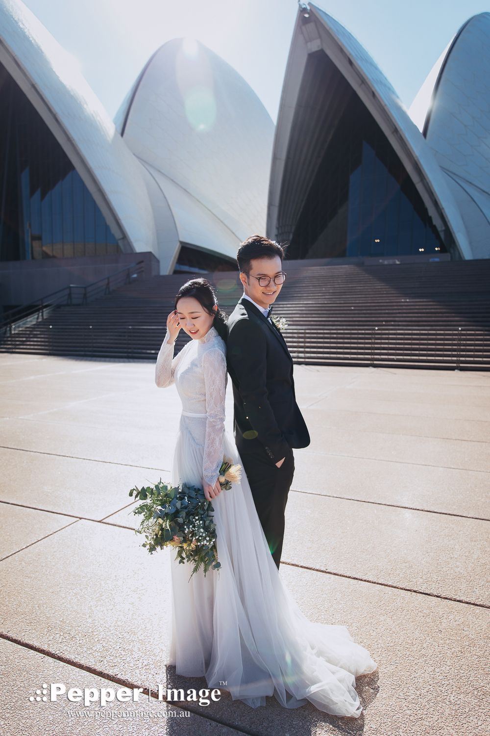 Bride holding bouquet and groom standing back to back at Sydney Opera House steps.
