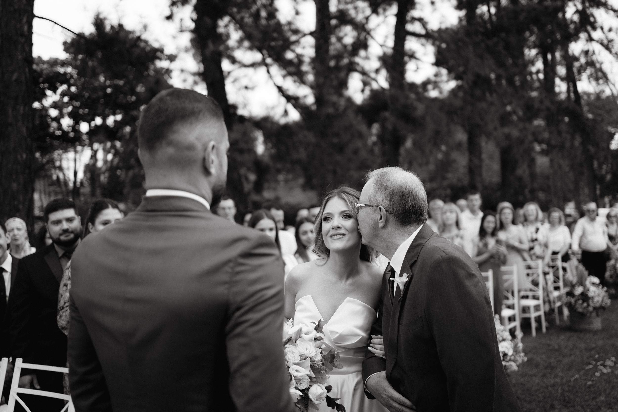 Wedding ceremony guests watch attentively from white chairs during outdoor celebration.