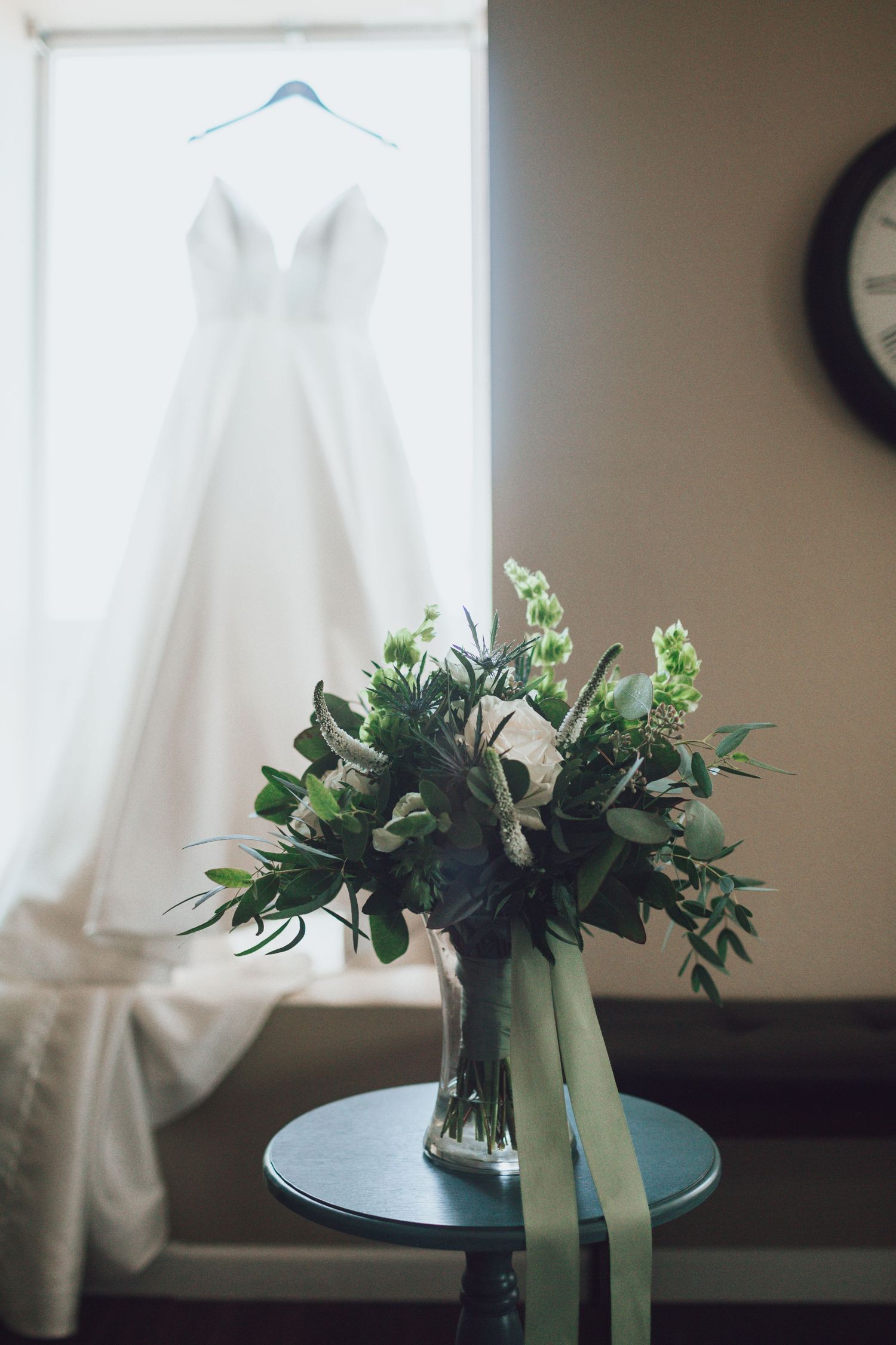 A white wedding dress hangs by a window with a floral bouquet displayed on a table below.
