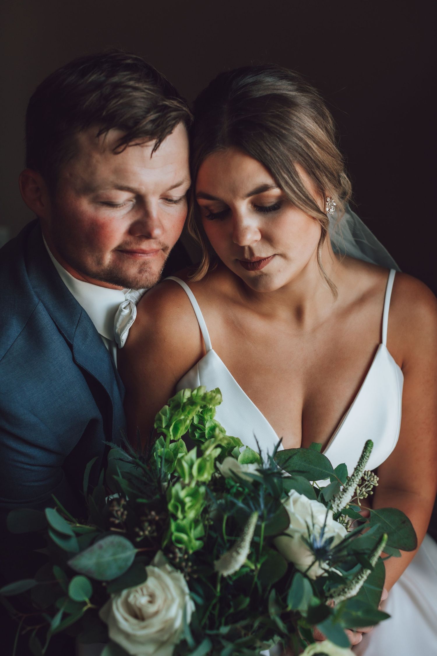 Close-up wedding portrait with white dress and greenery bouquet in dramatic lighting.