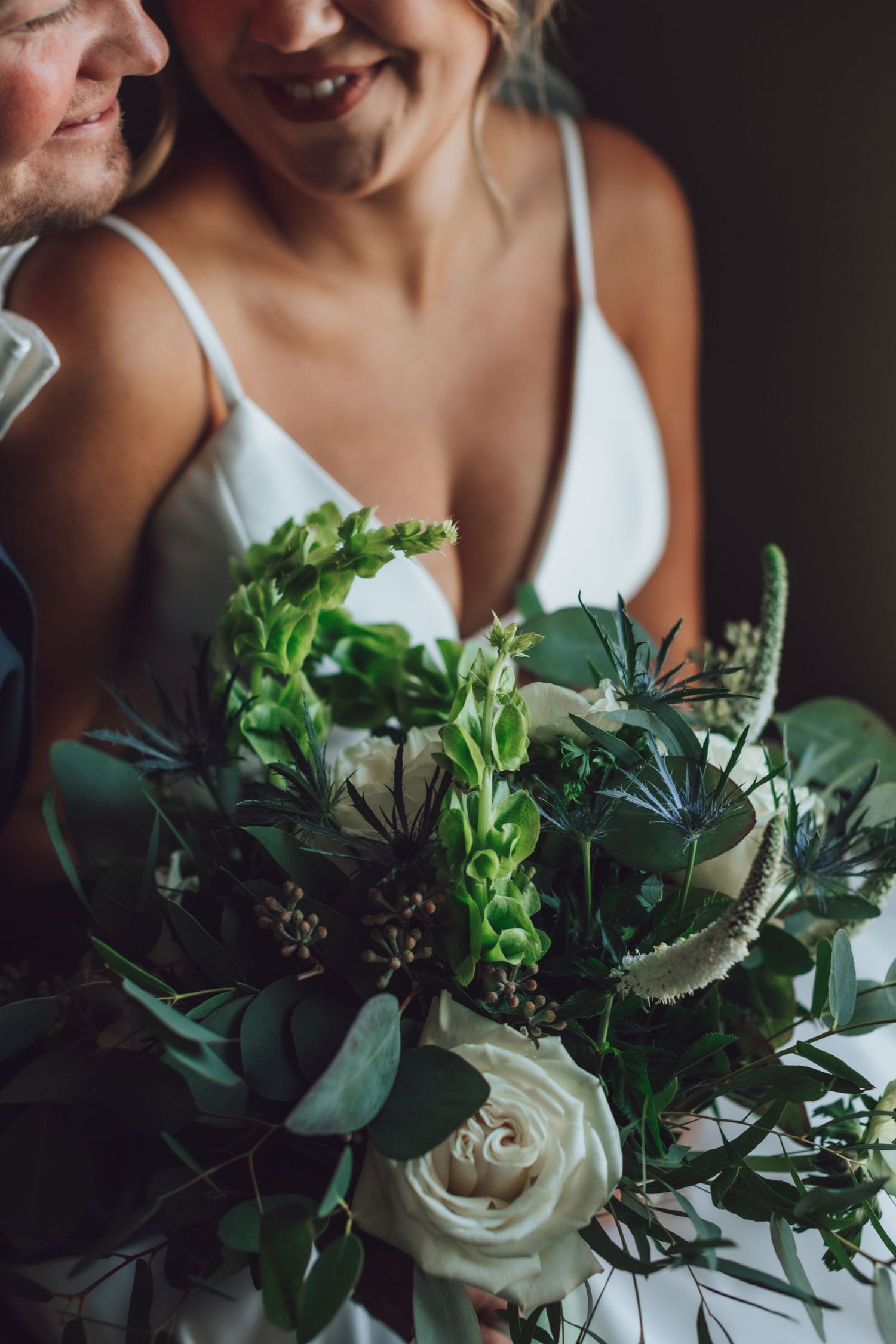 Wedding bouquet features white roses and lush greenery in moody lighting.