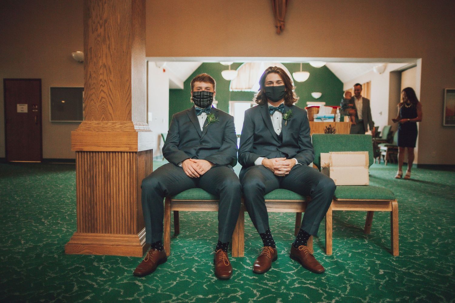 Groomsmen in dark suits sit casually on church bench with green carpet.