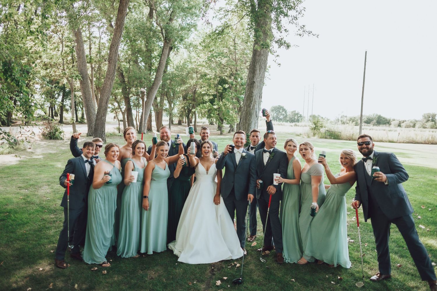 Wedding party members strike fun poses together on a grassy lawn with trees in background.