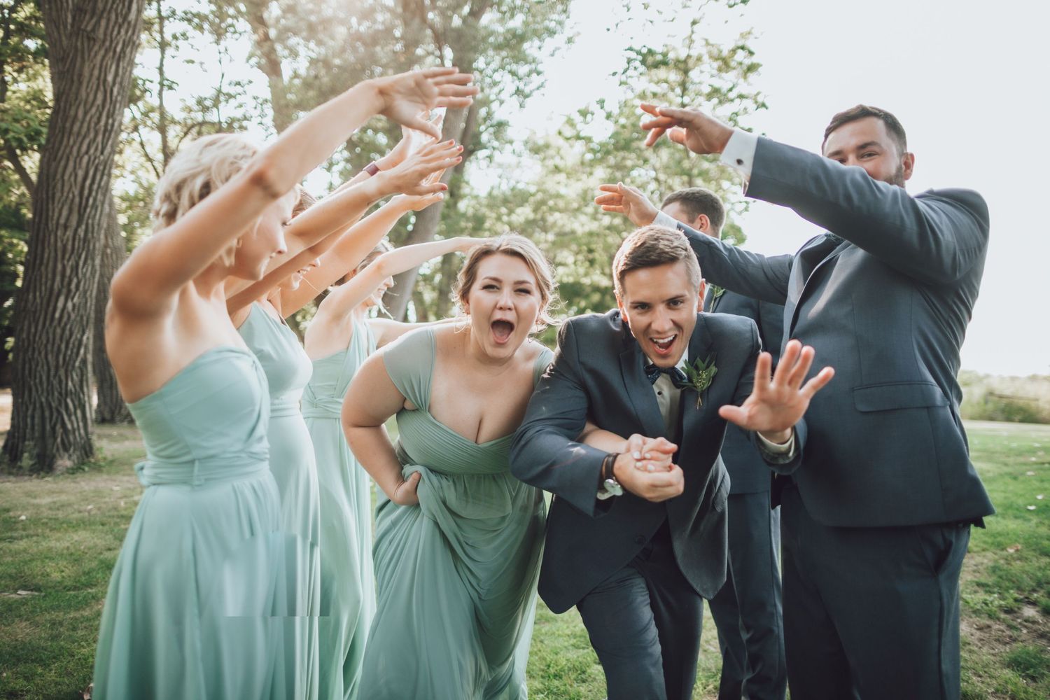 Bridal party jumps and poses energetically for photos outdoors on a sunny day.