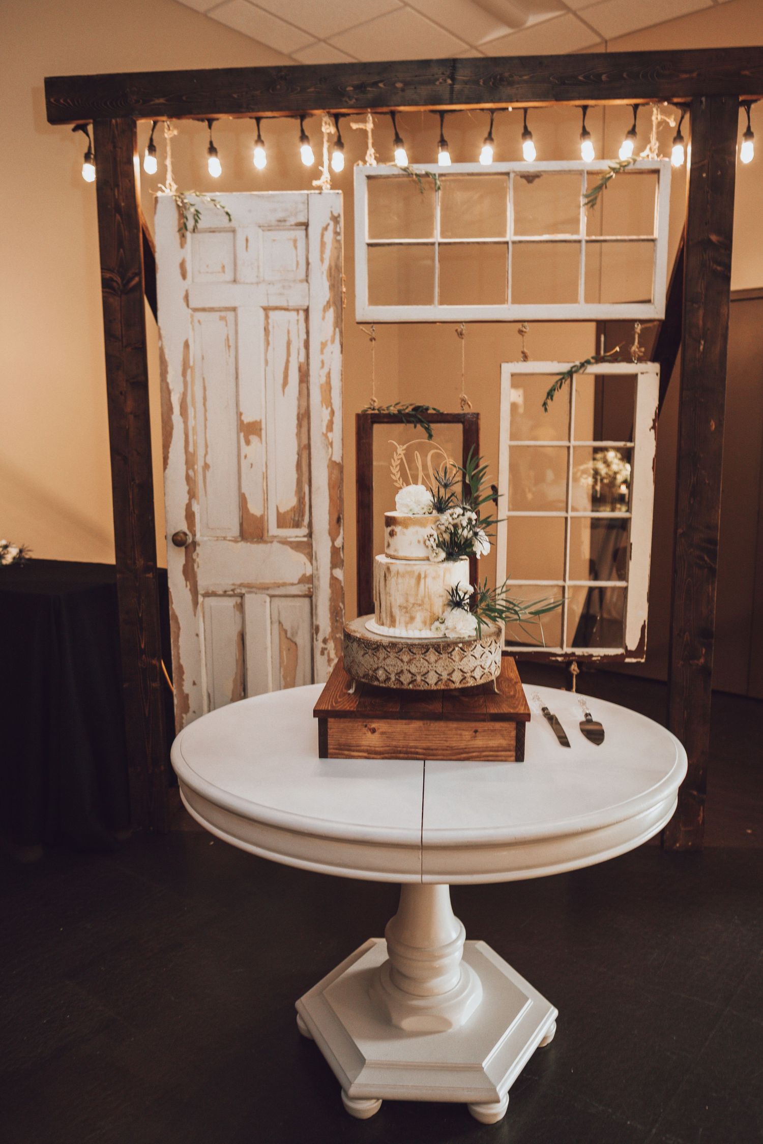 Elegant white wedding cake displayed on pedestal table with rustic barn door backdrop and string lights.