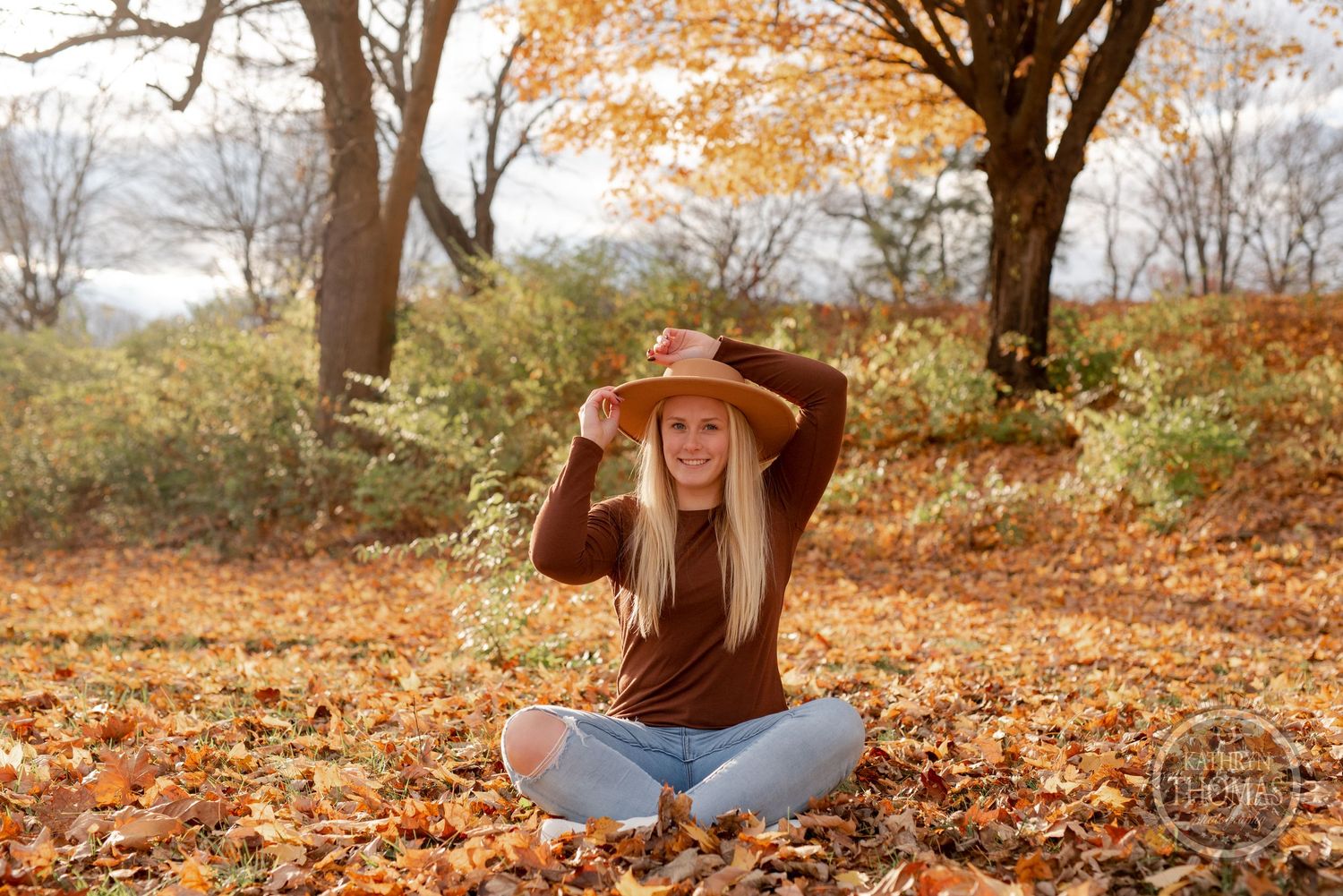 Person in brown sweater sits cross-legged on fallen leaves under autumn trees.