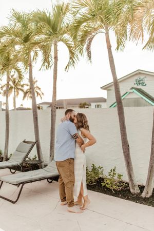 A couple shares an intimate embrace on a tropical patio with tall palm trees and lounge chairs.