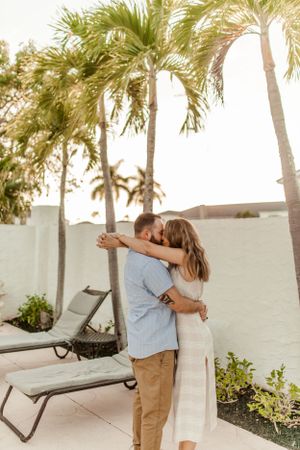 A couple shares a tender moment while hugging on a resort patio under palm trees.