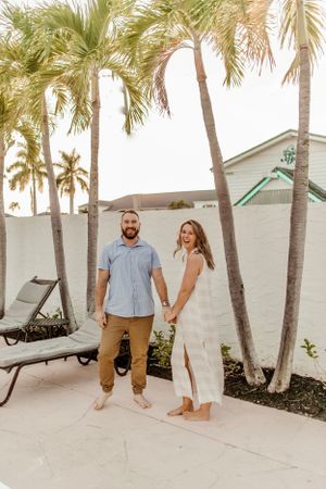 A couple stands together on a tropical patio with palm trees and lounge chairs.