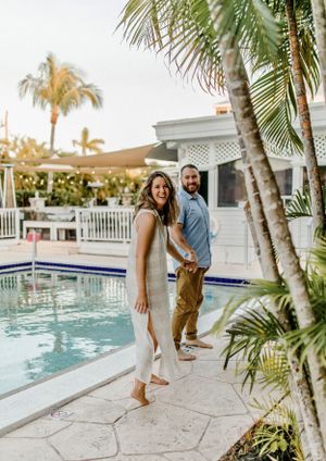 Couple poses playfully by a swimming pool surrounded by palm trees and white architecture.