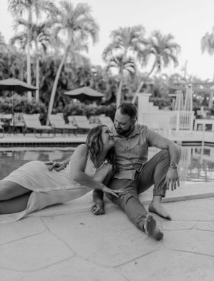 Black and white photo of couple lounging by pool with palm trees in background.