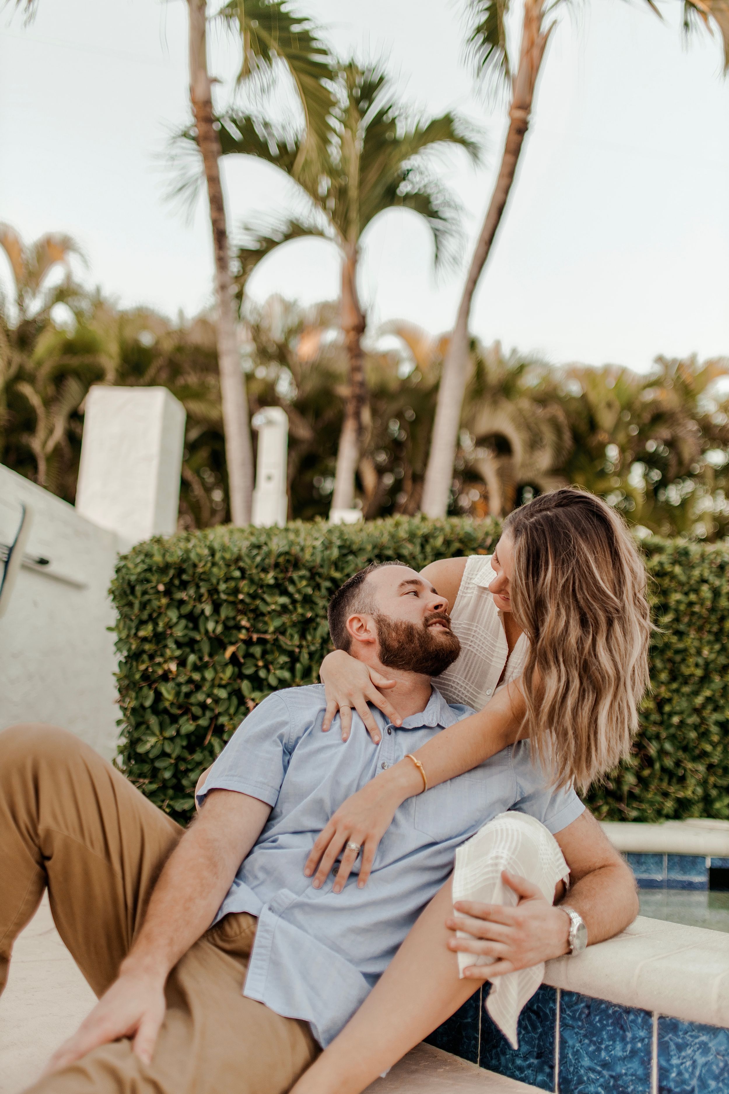 Couple sharing tender moment while sitting poolside with tropical garden backdrop.