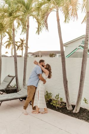 A romantic couple embraces under palm trees on a tropical patio near lounge chairs.