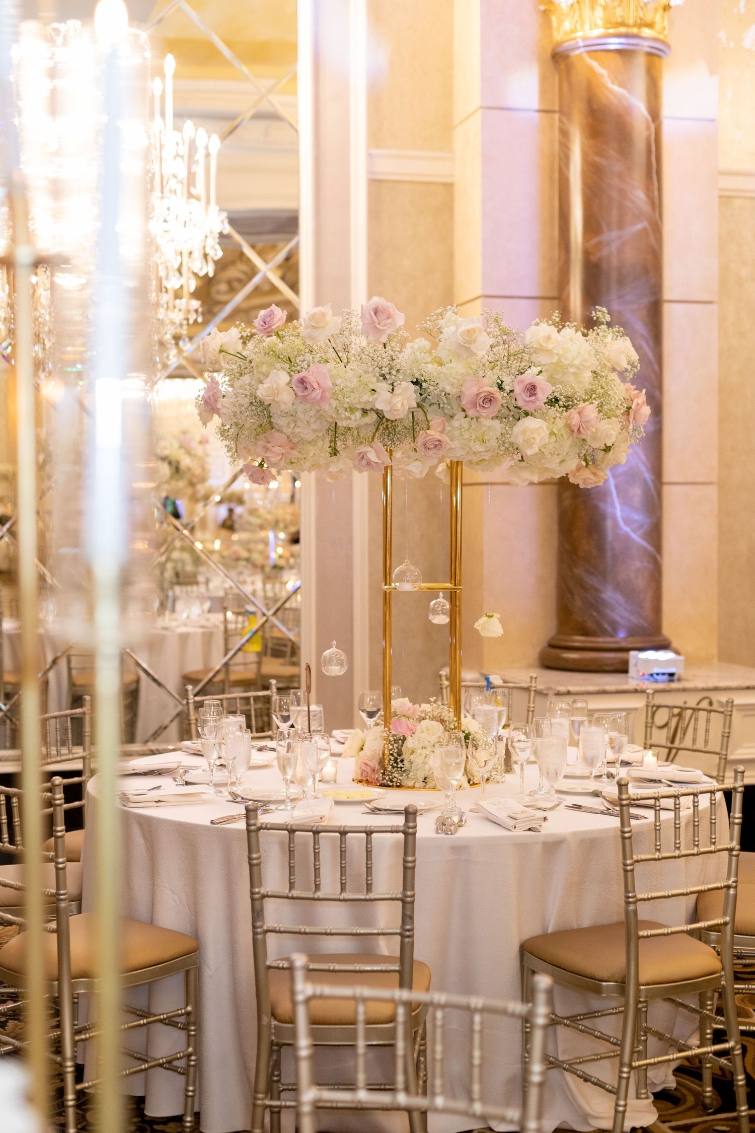Elegant wedding reception table with tall floral centerpiece, crystal chandelier, and gold chiavari chairs.