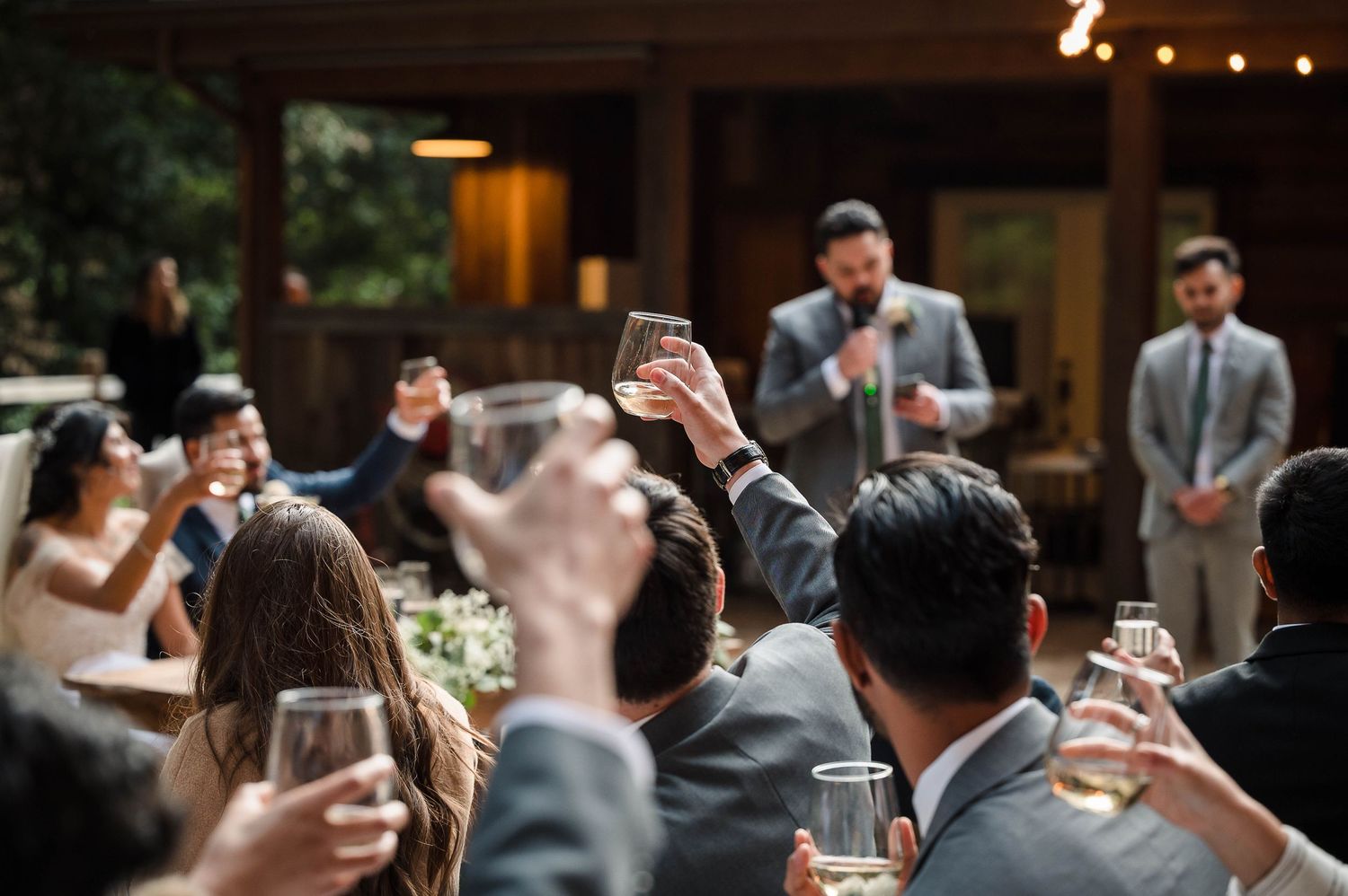Wedding guests raise their glasses in a toast during an outdoor reception with string lights overhead.