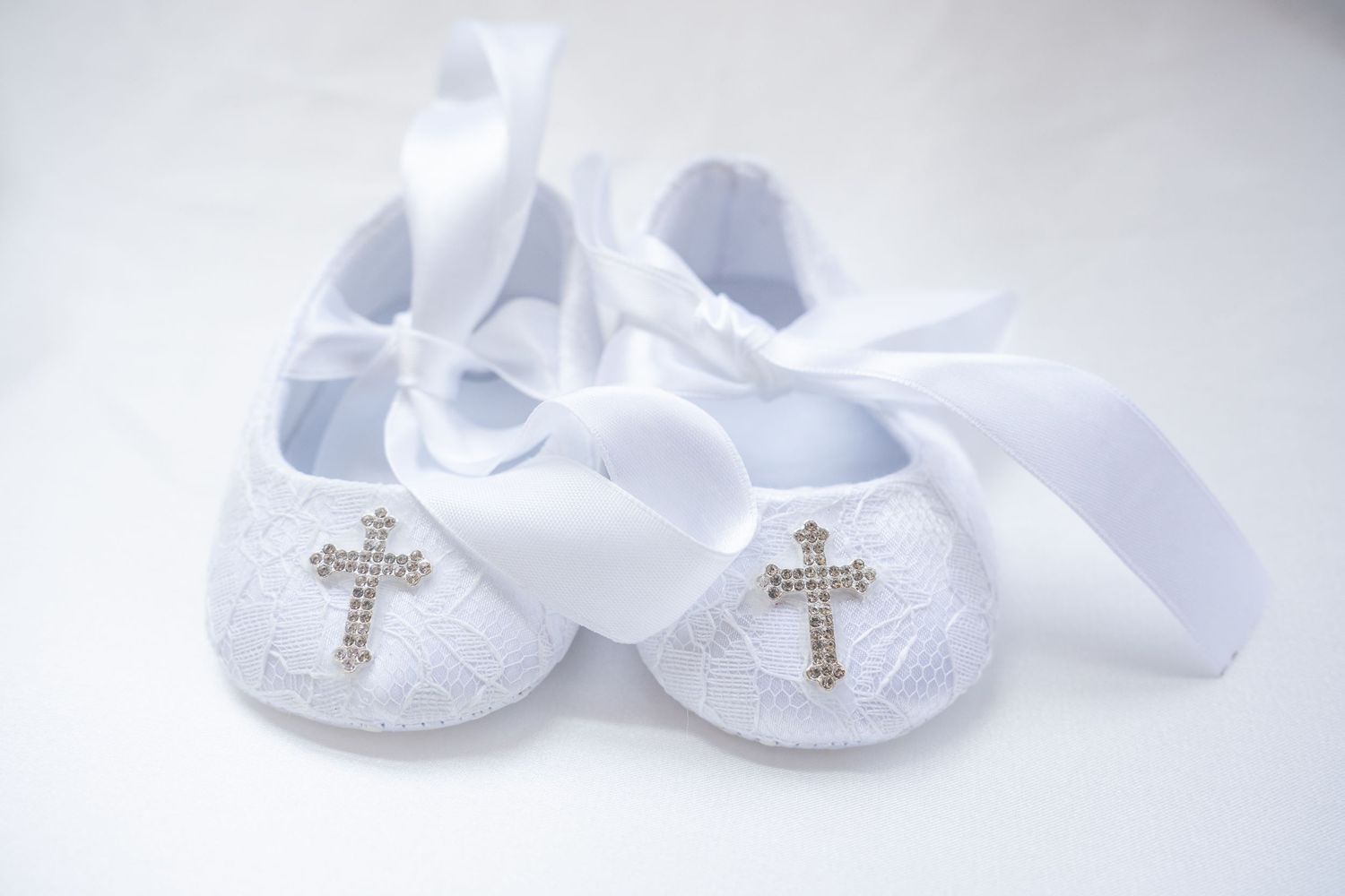 White baby shoes with delicate cross details and ribbons on a light background.