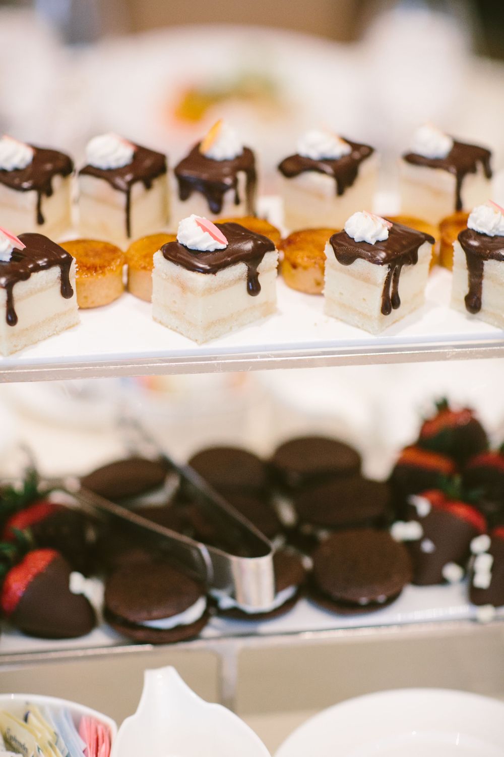 Elegant dessert display featuring chocolate-drizzled petit fours and chocolate-covered strawberries on tiered serving plates.