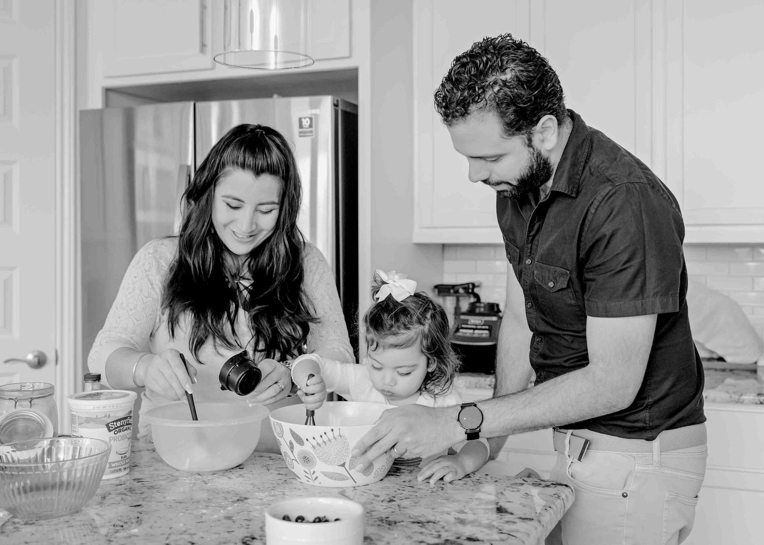 A family bakes together in a bright white kitchen, working with ingredients on a flour-dusted counter.