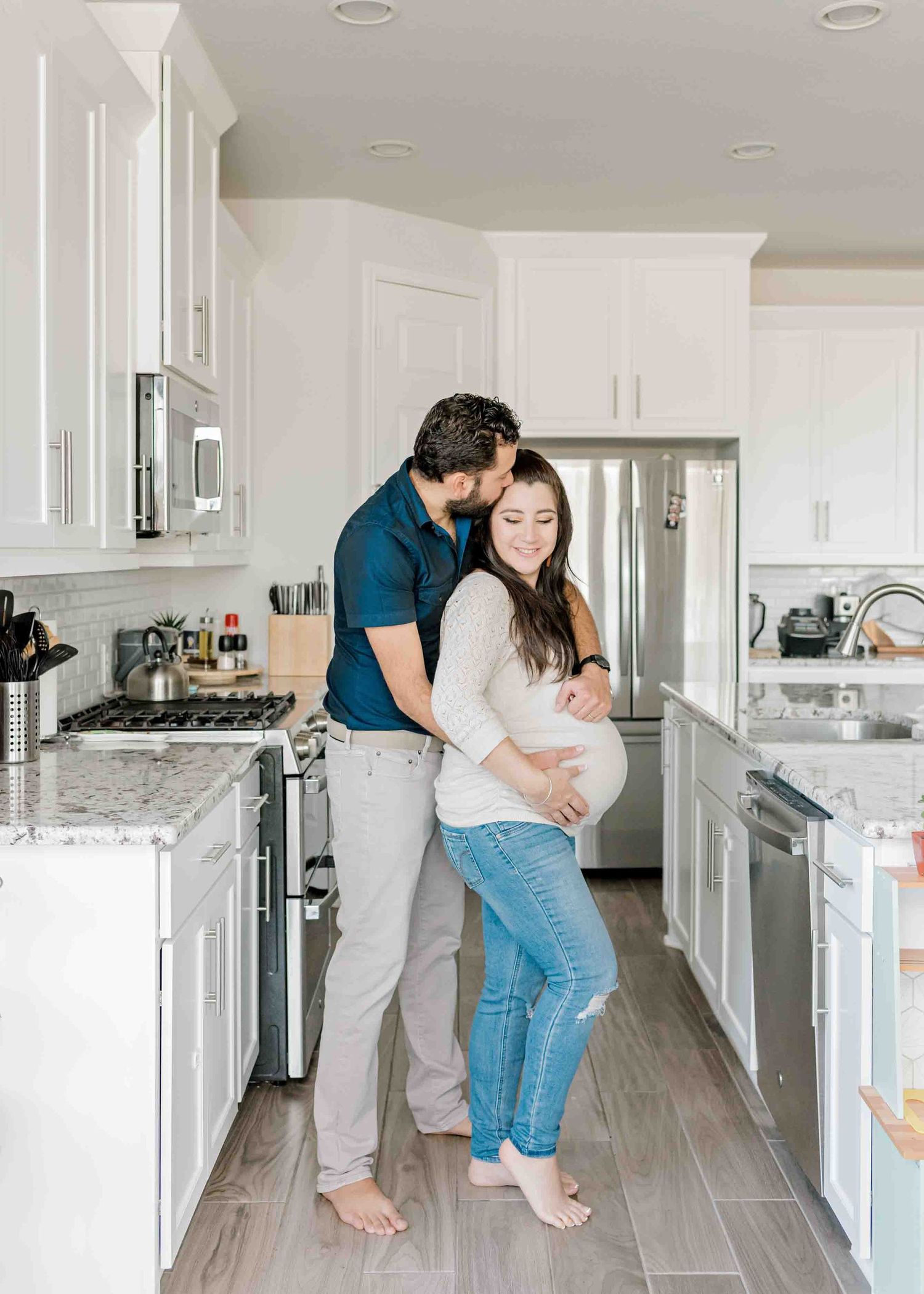 A couple embraces in their modern white kitchen with hardwood floors.