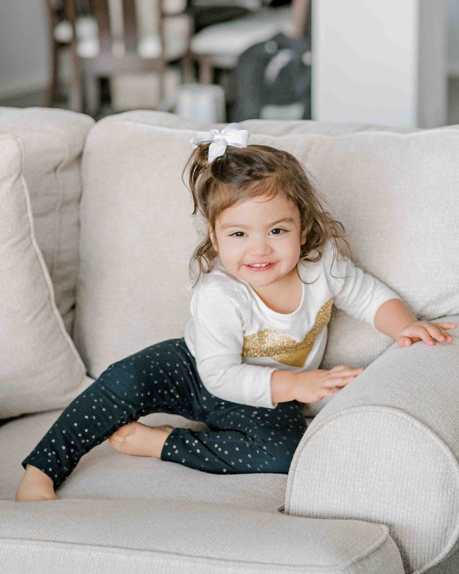 A toddler in a sparkly outfit relaxes on a plush cream-colored couch.