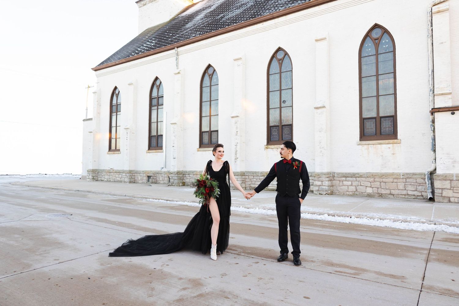 Elegant couple in formal attire poses outside a white church with arched windows.