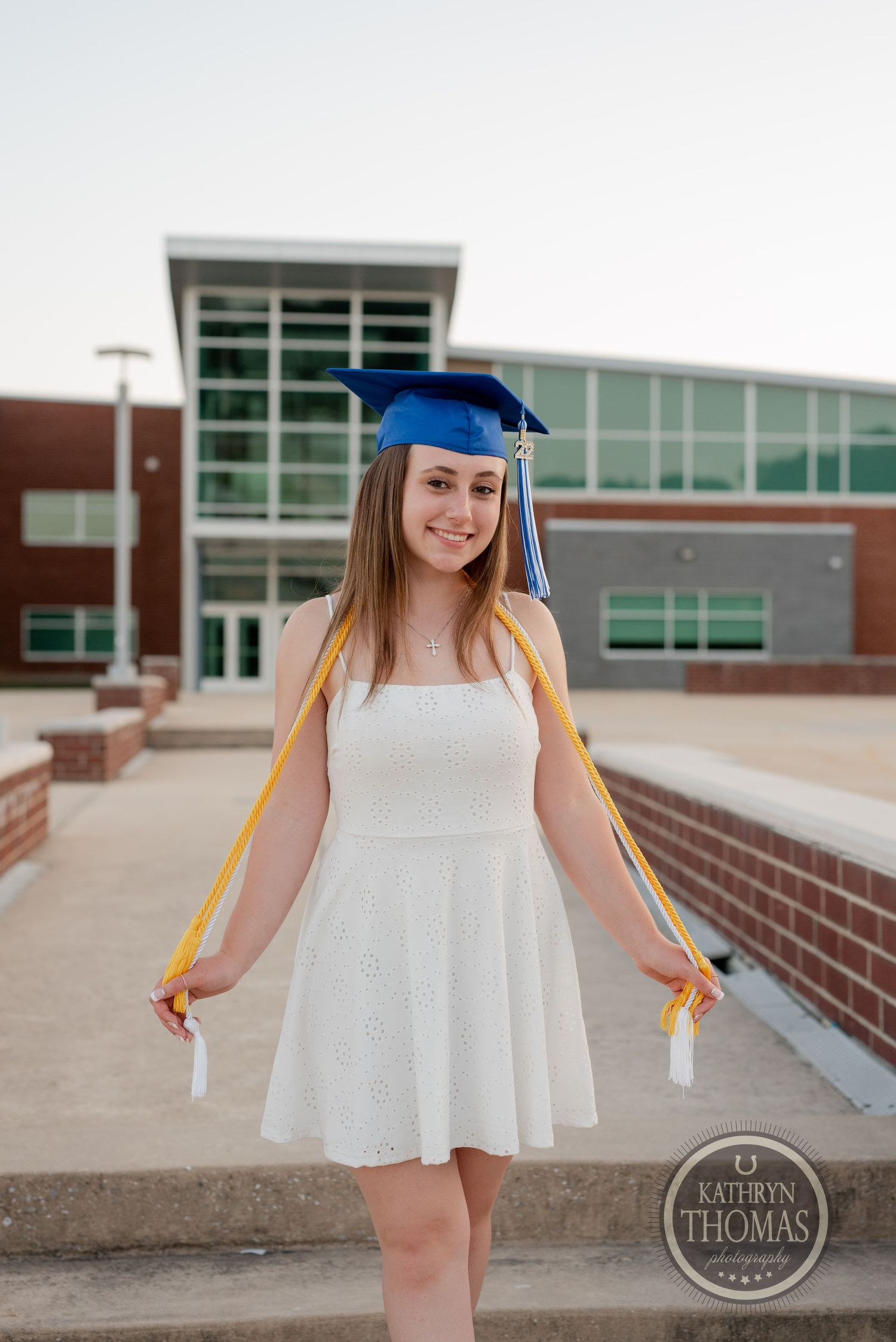 Graduate in white dress and blue regalia poses with yellow honor cords on campus.