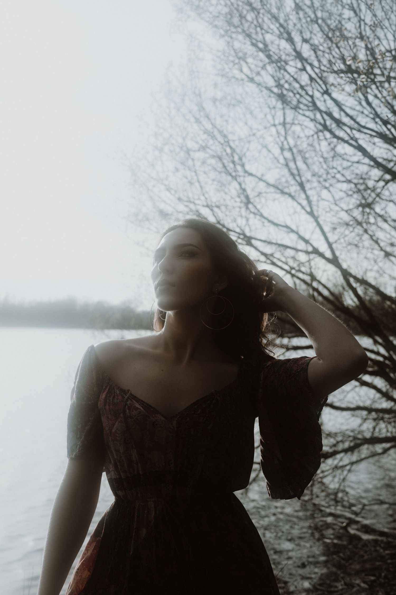 Atmospheric portrait of woman in dark dress by misty lake with bare trees.