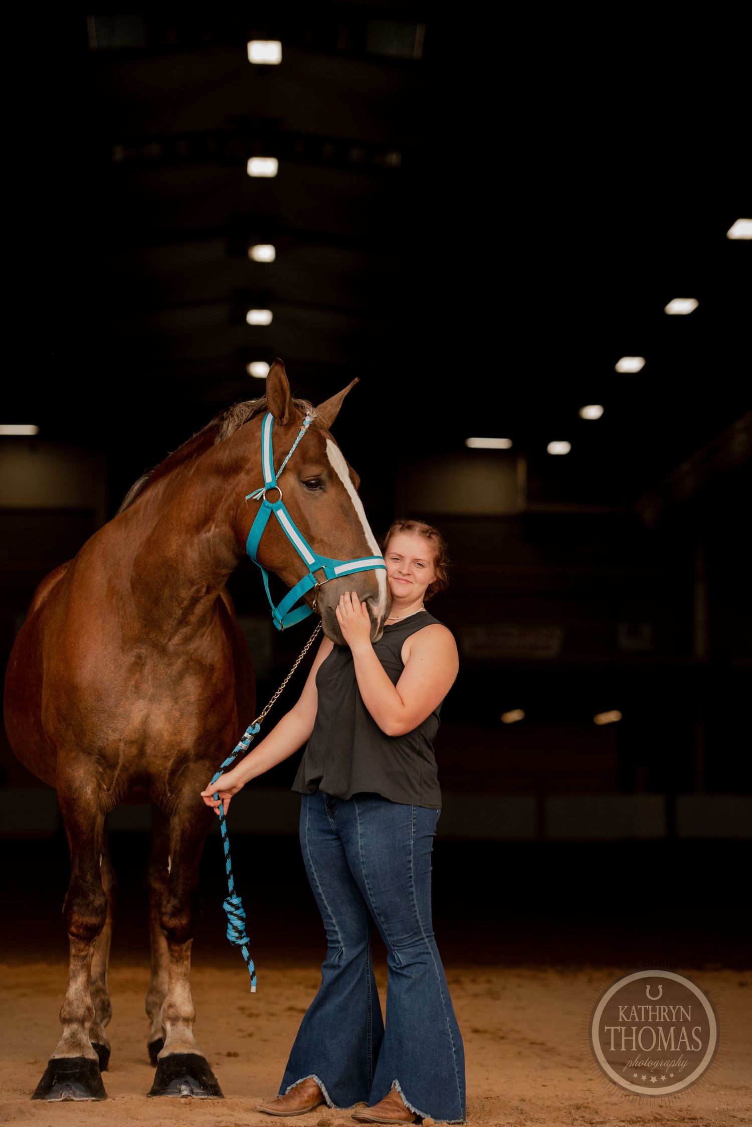 A dark horse with turquoise tack stands with its handler in an indoor riding arena.