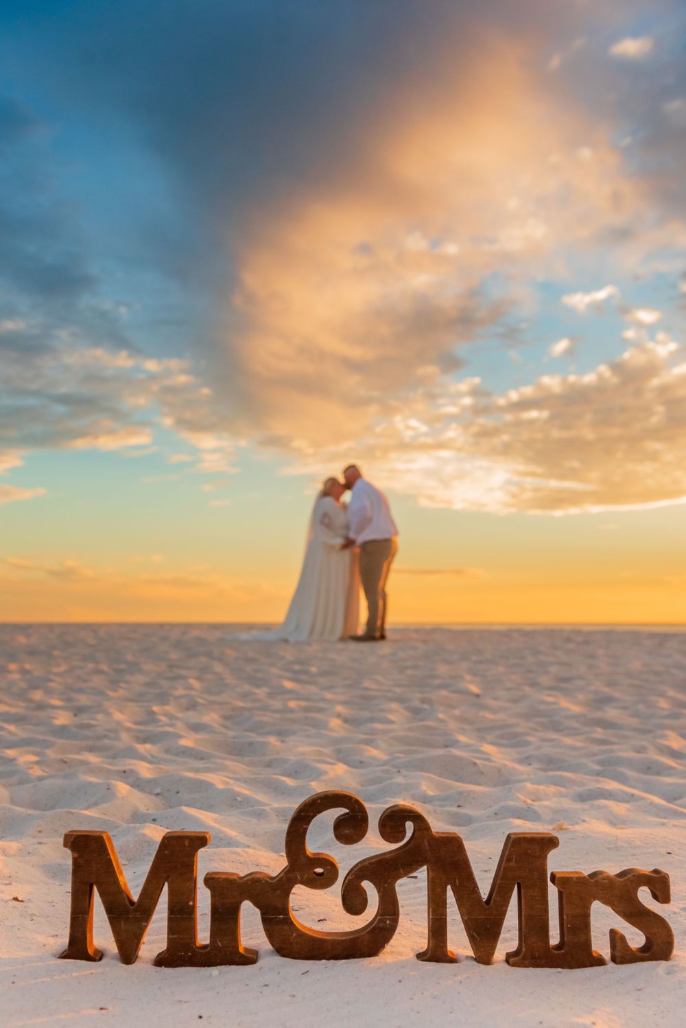 mr and mrs sign in foreground with bride & groom kissing at sunset on their wedding day on Panama City Beach Florida