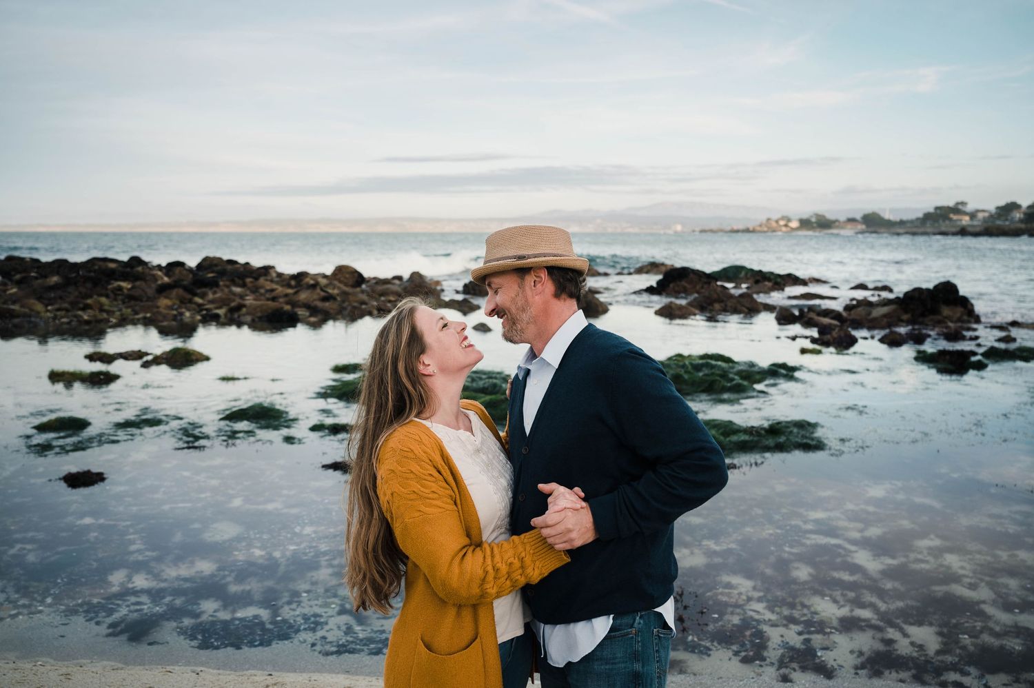 Couple sharing intimate moment on coastal beach with rocks and ocean backdrop.