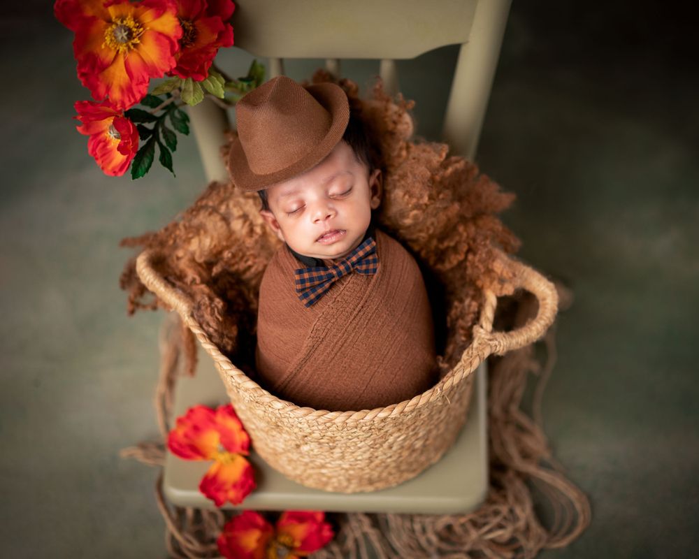 A sleeping newborn wearing brown clothing and bow tie lies in a woven basket with red floral accents.