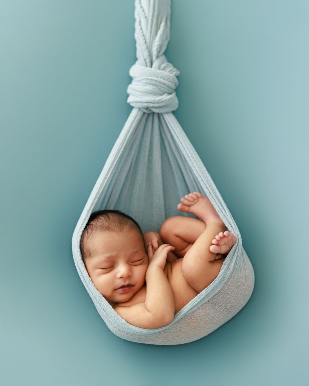 Peaceful newborn sleeping in suspended white fabric hammock against blue background.