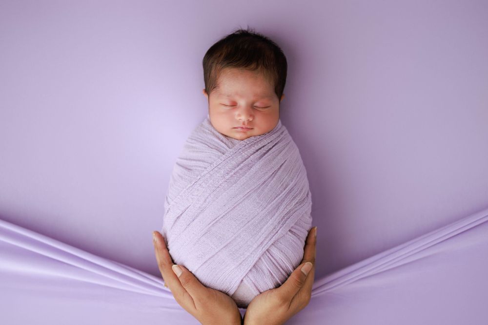 Sleeping newborn swaddled in white fabric against purple background.