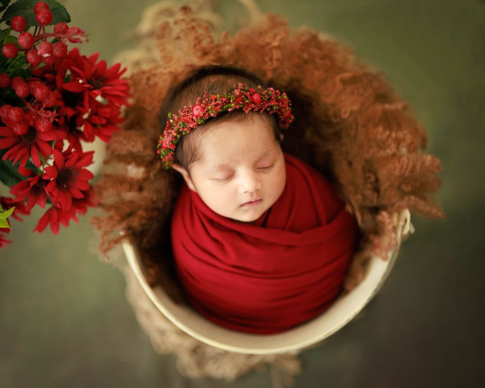 Peaceful newborn wrapped in red fabric with floral crown near vibrant red flowers.