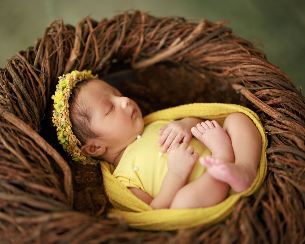Sleeping newborn wrapped in yellow fabric nesting in brown woven basket with floral headpiece.