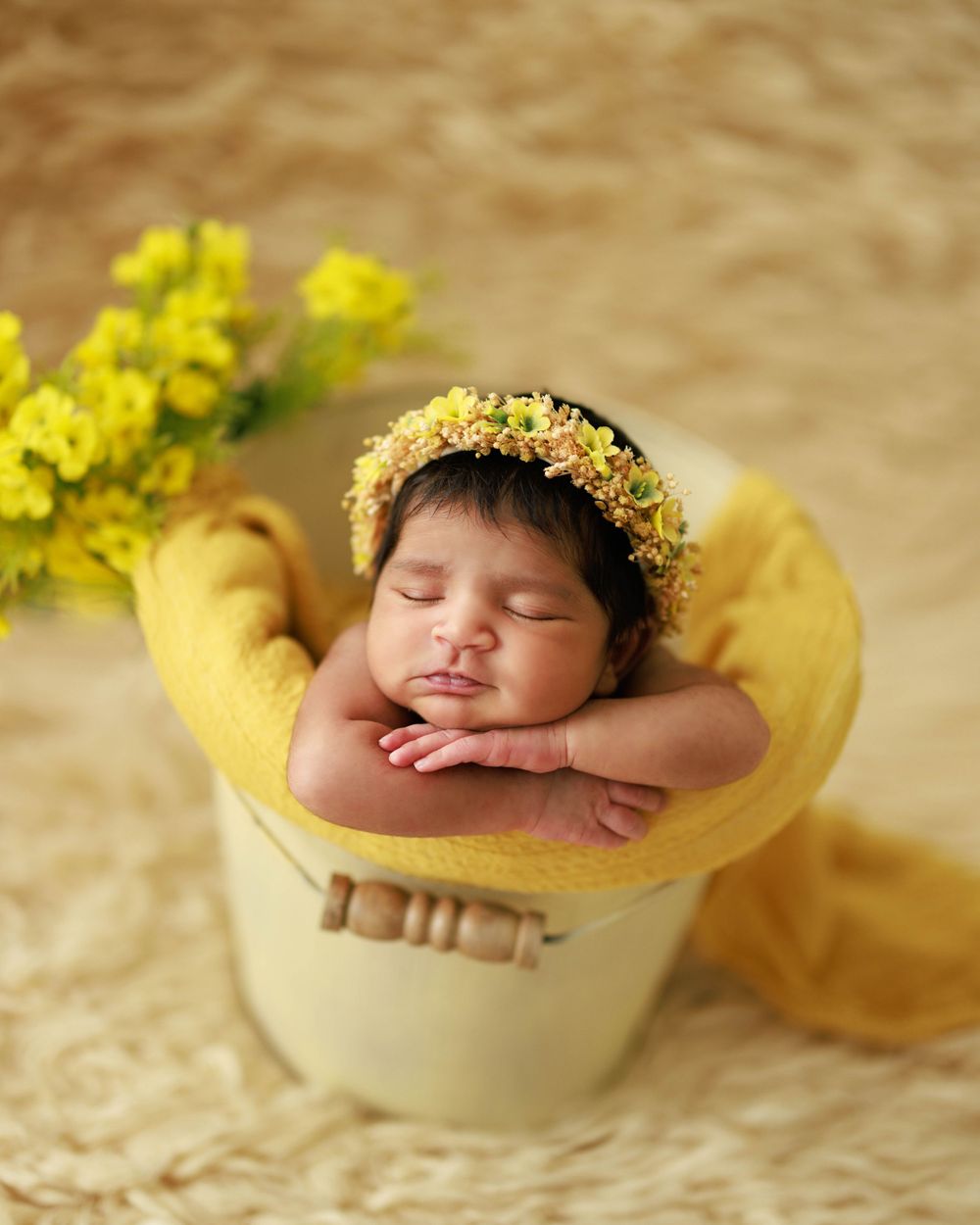 Sleeping newborn with yellow floral crown resting in cream-colored container.