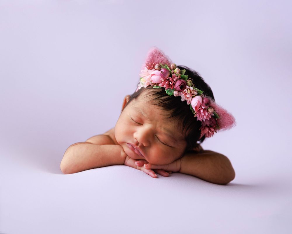 Sleeping newborn wearing floral crown headpiece on soft purple background.