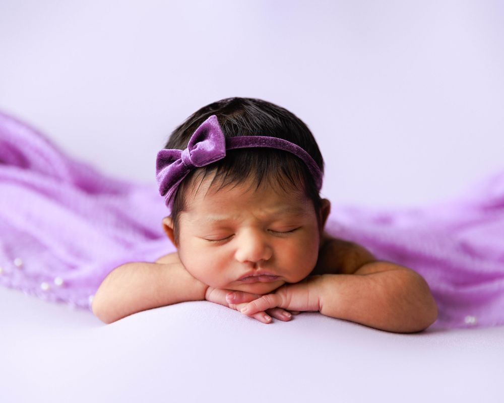 Peaceful sleeping newborn on purple backdrop wearing matching purple bow headband.