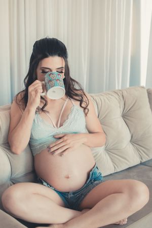 Casual maternity moment on grey couch drinking from blue mug in lace bralette.