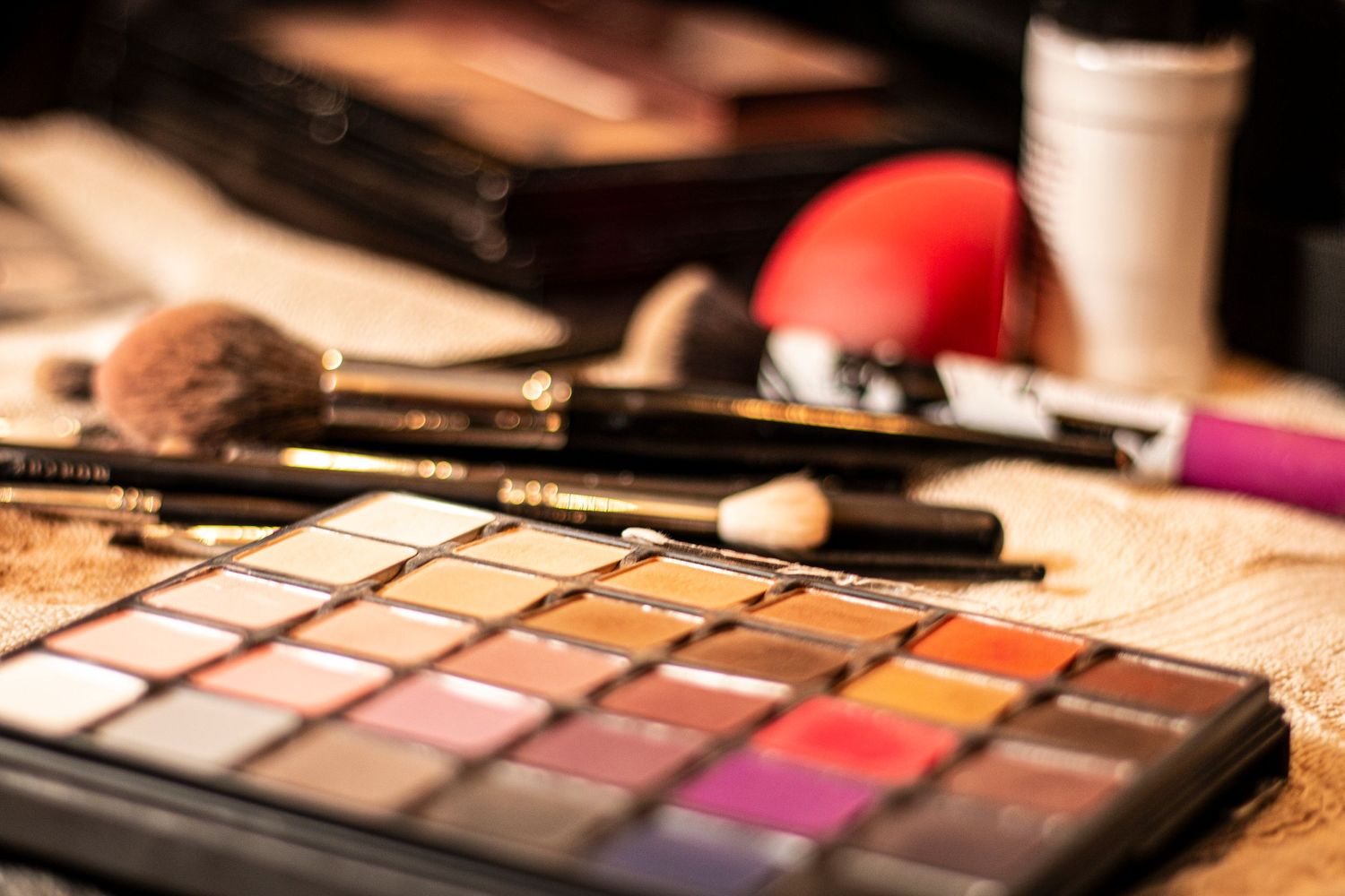 Closeup of a makeup palette with eyeshadow colors alongside brushes and beauty accessories on a textured surface.