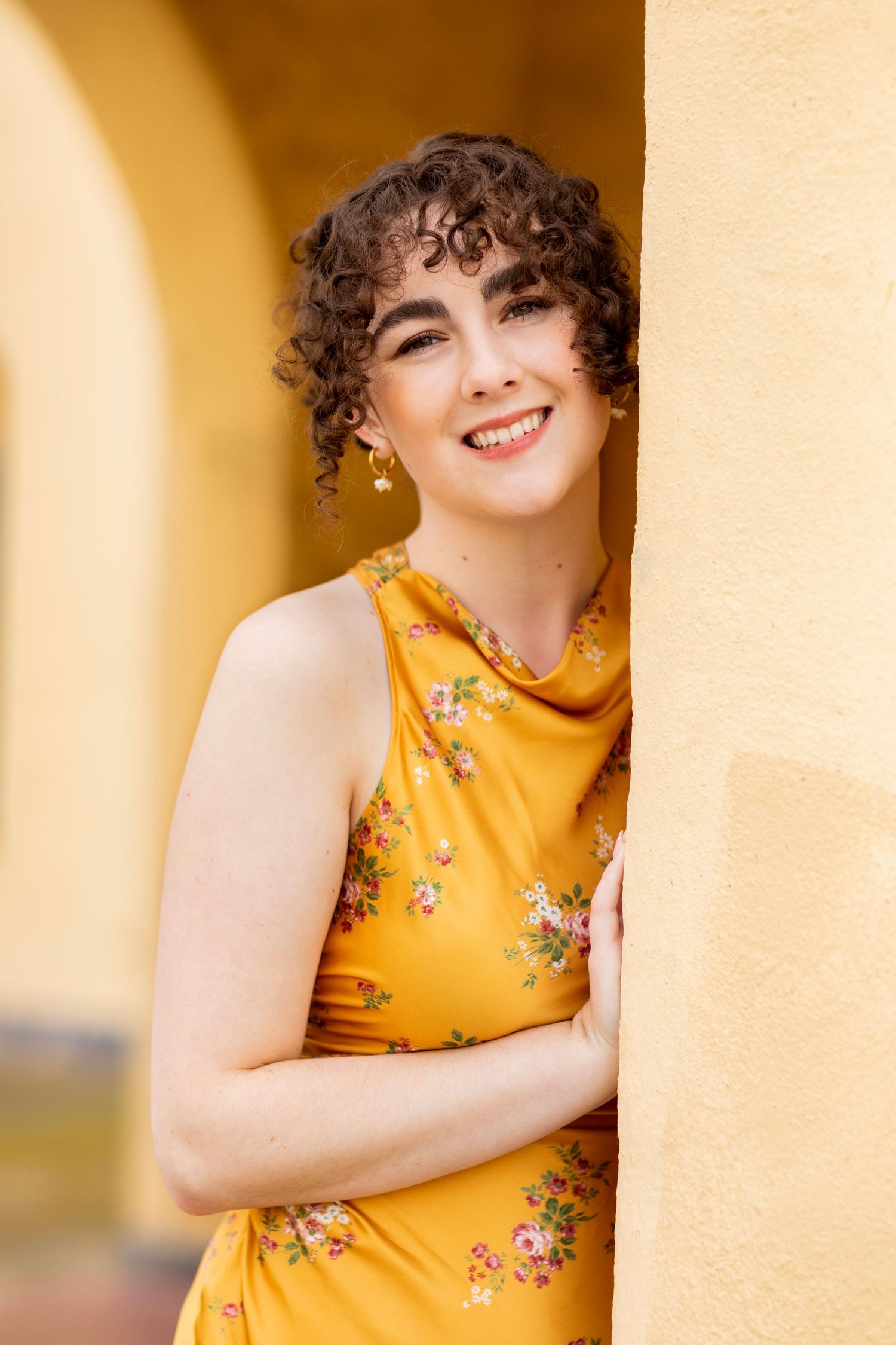 A high school senior with curly hair wearing a yellow floral dress poses against an archway in warm lighting.