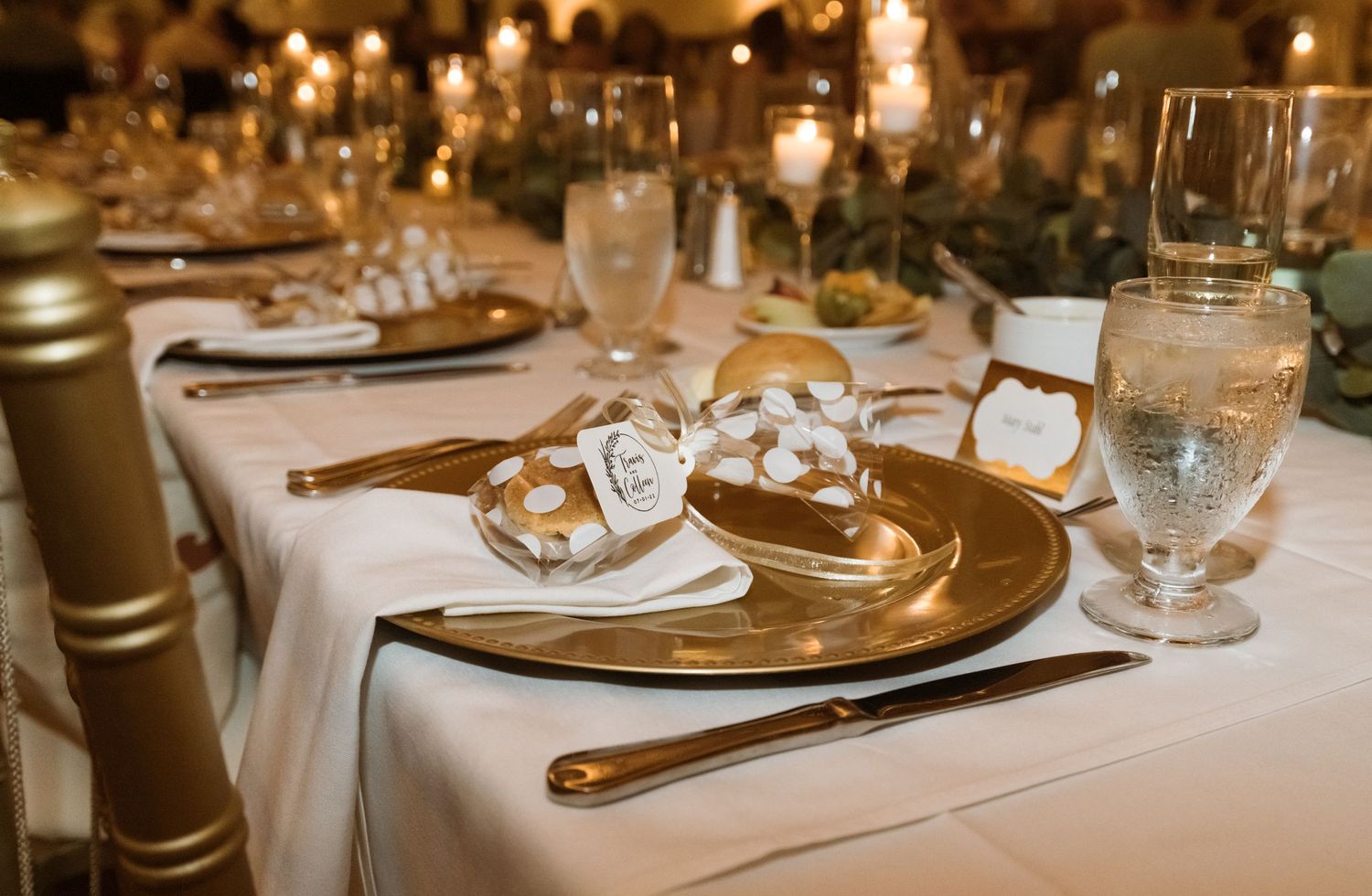 Elegant wedding reception table setting with gold charger plates and crystal glasses on white linens.