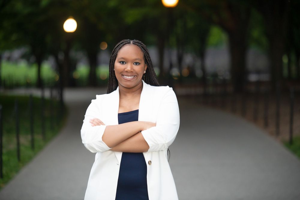 Graduate in white blazer and dark dress poses outdoors with soft lighting in the background.