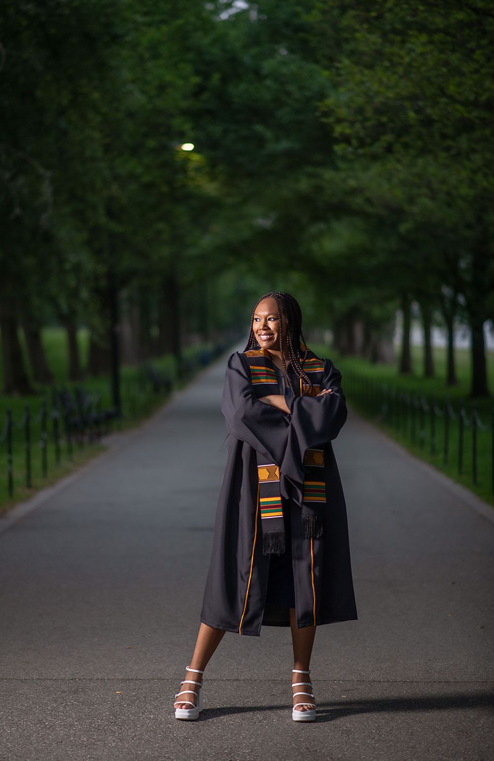 Graduate in black gown and stole poses playfully on tree-lined path during evening photo session.