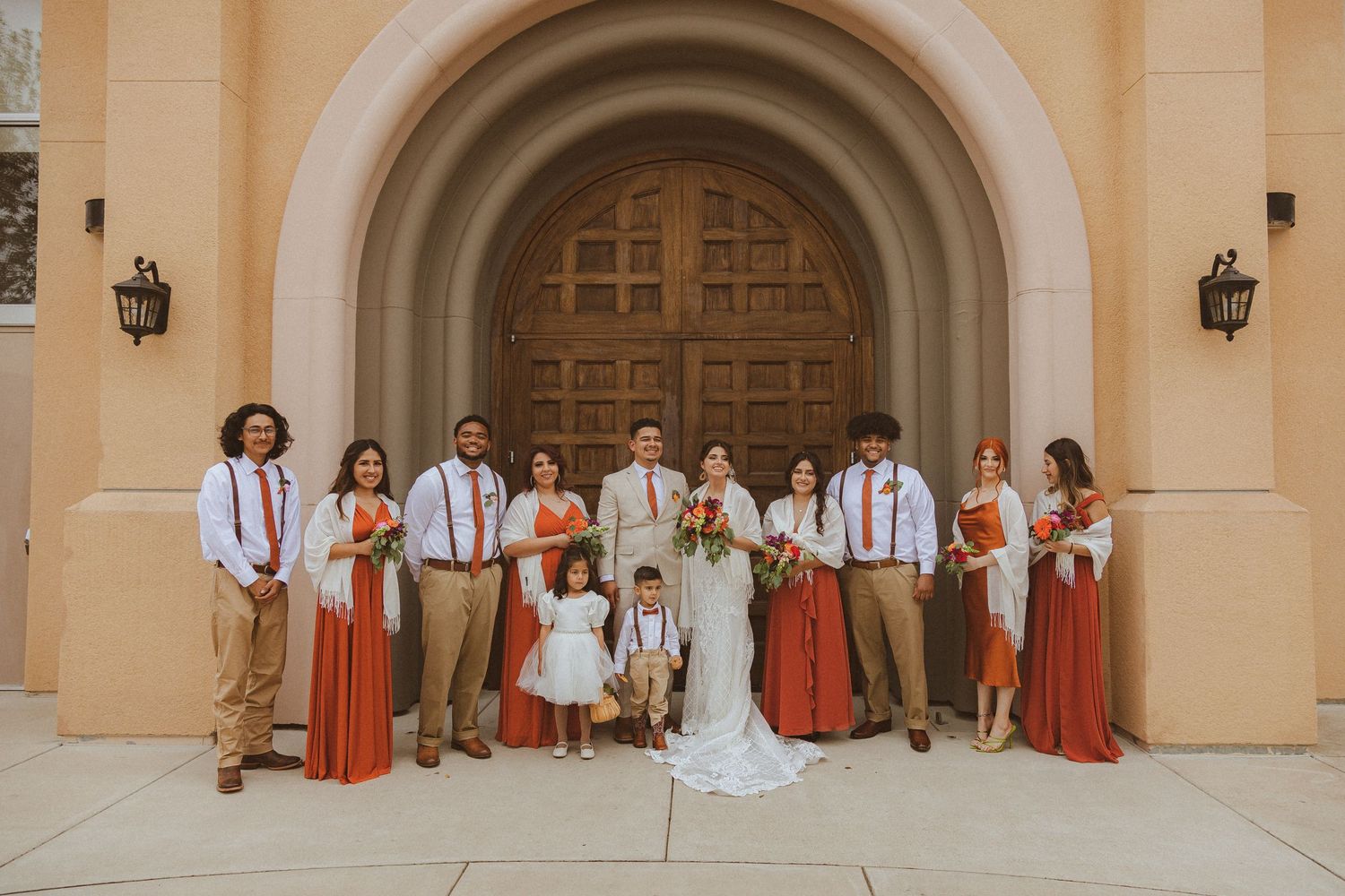 Wedding party in coral dresses and khaki attire poses together in front of church entrance.